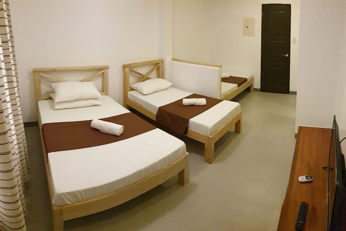A hotel room with three wooden beds, each with white linens and brown bedspreads. There is a wooden desk with a television and remote controls.