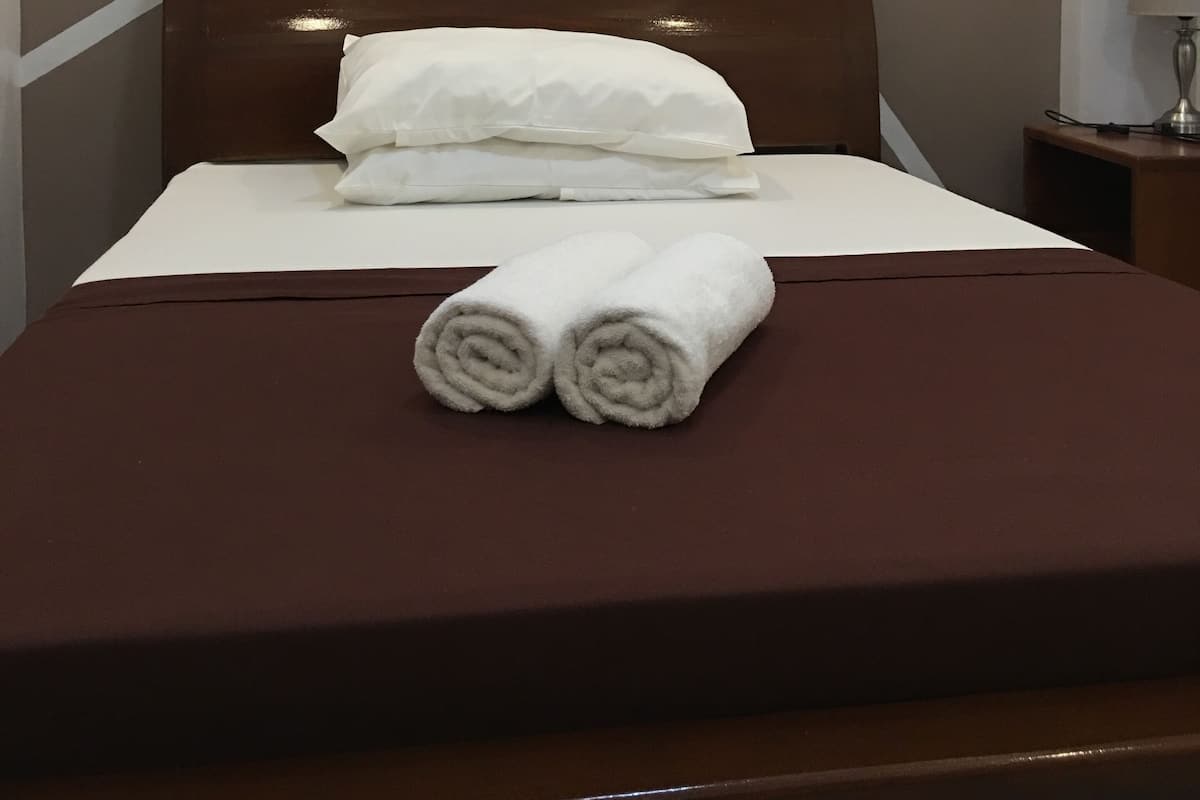 A neatly made bed with white linens and two rolled towels, a wooden headboard, and a wall with a geometric pattern.