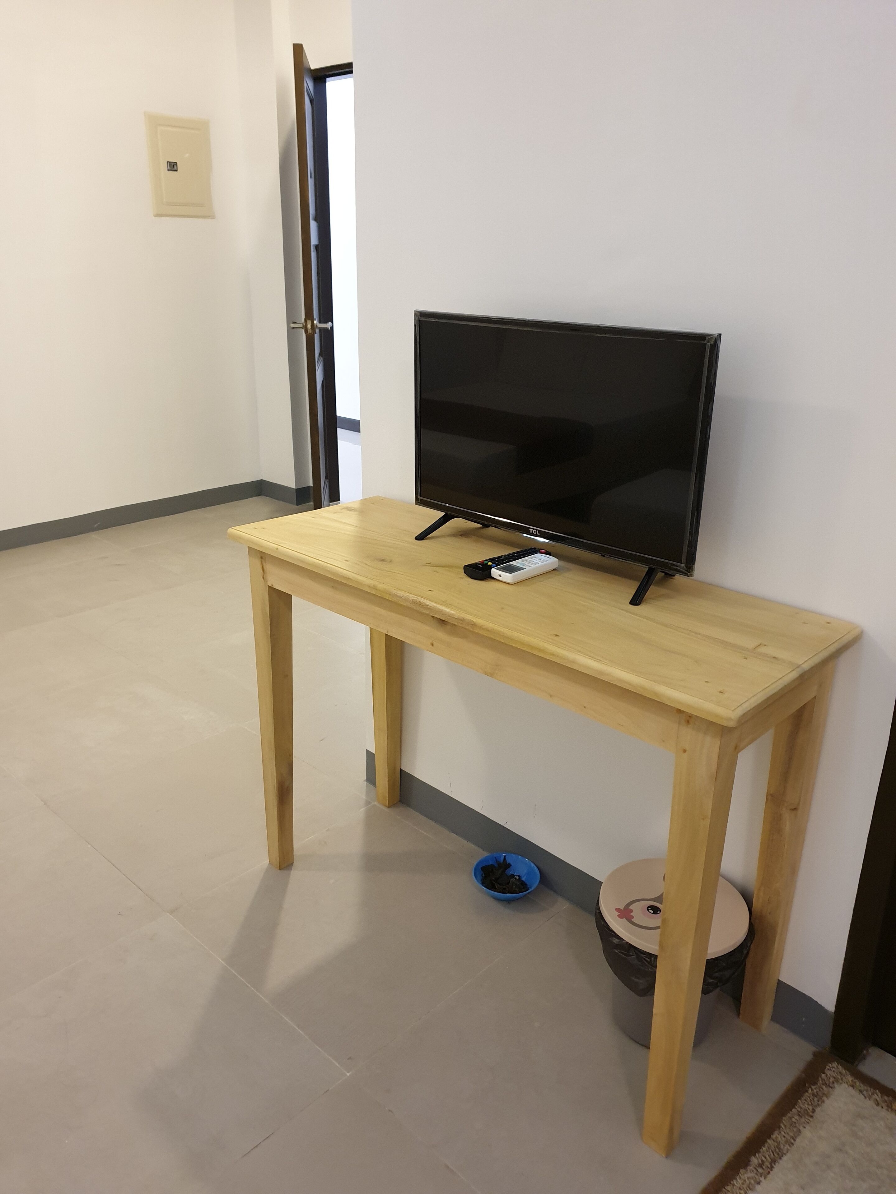 A wooden TV stand with a flat-screen TV and remote control, a small blue bowl, and a trash can with a lid.