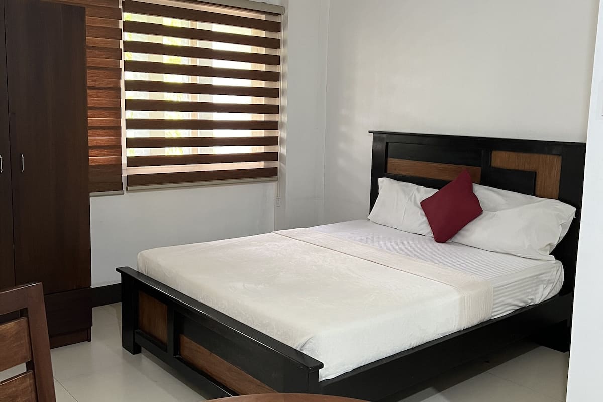 A bedroom with a bed, a wooden wardrobe, a window with blinds, and a round table.
