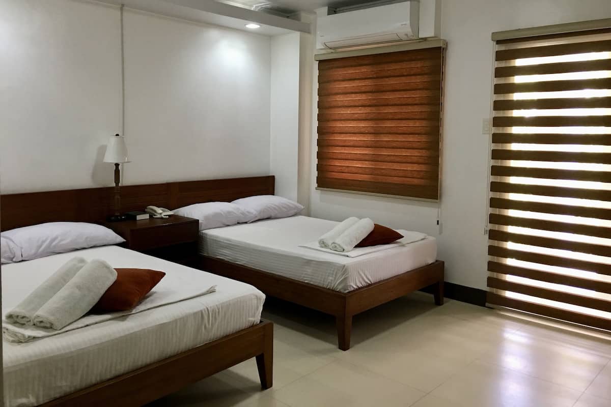 A hotel room with two single beds, a nightstand with a lamp, an air conditioning unit, and a window with blinds.