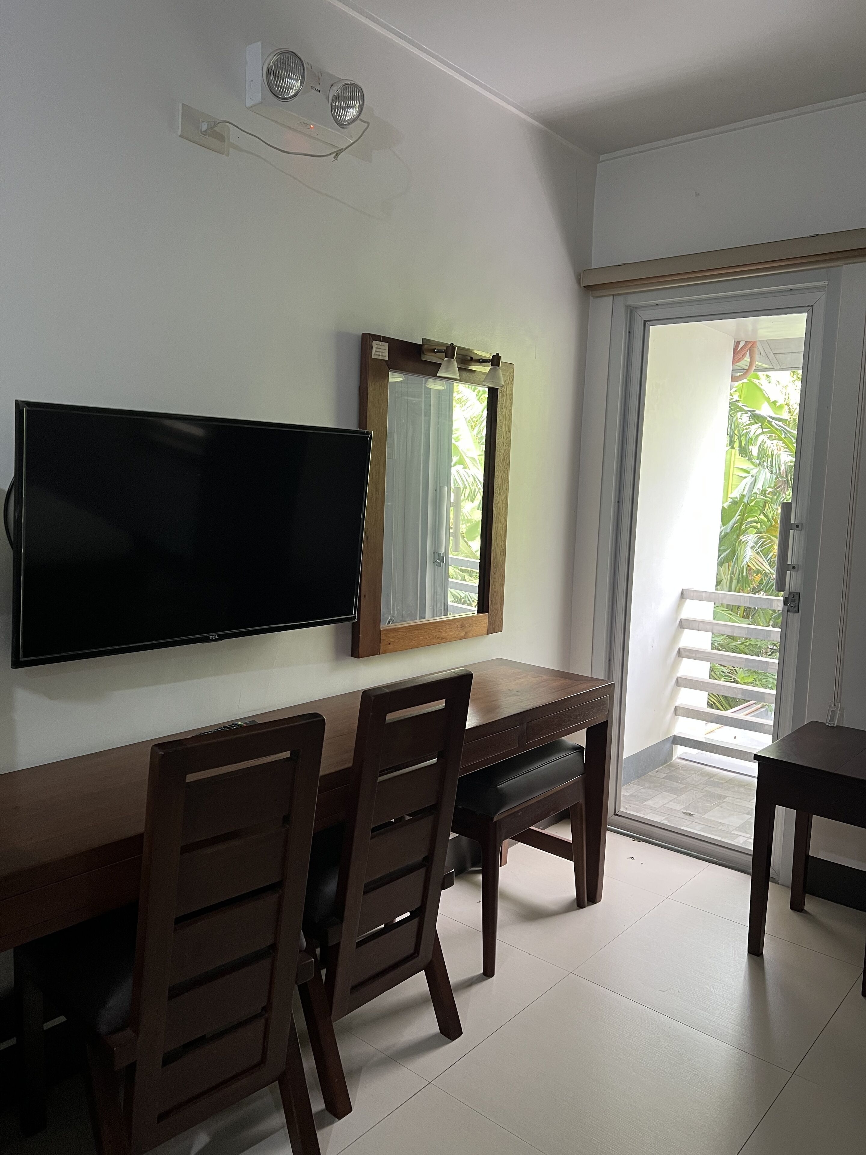 A room with a flat-screen TV mounted on the wall, a wooden desk with two chairs, a large mirror, and a sliding glass door leading to a balcony with greenery.