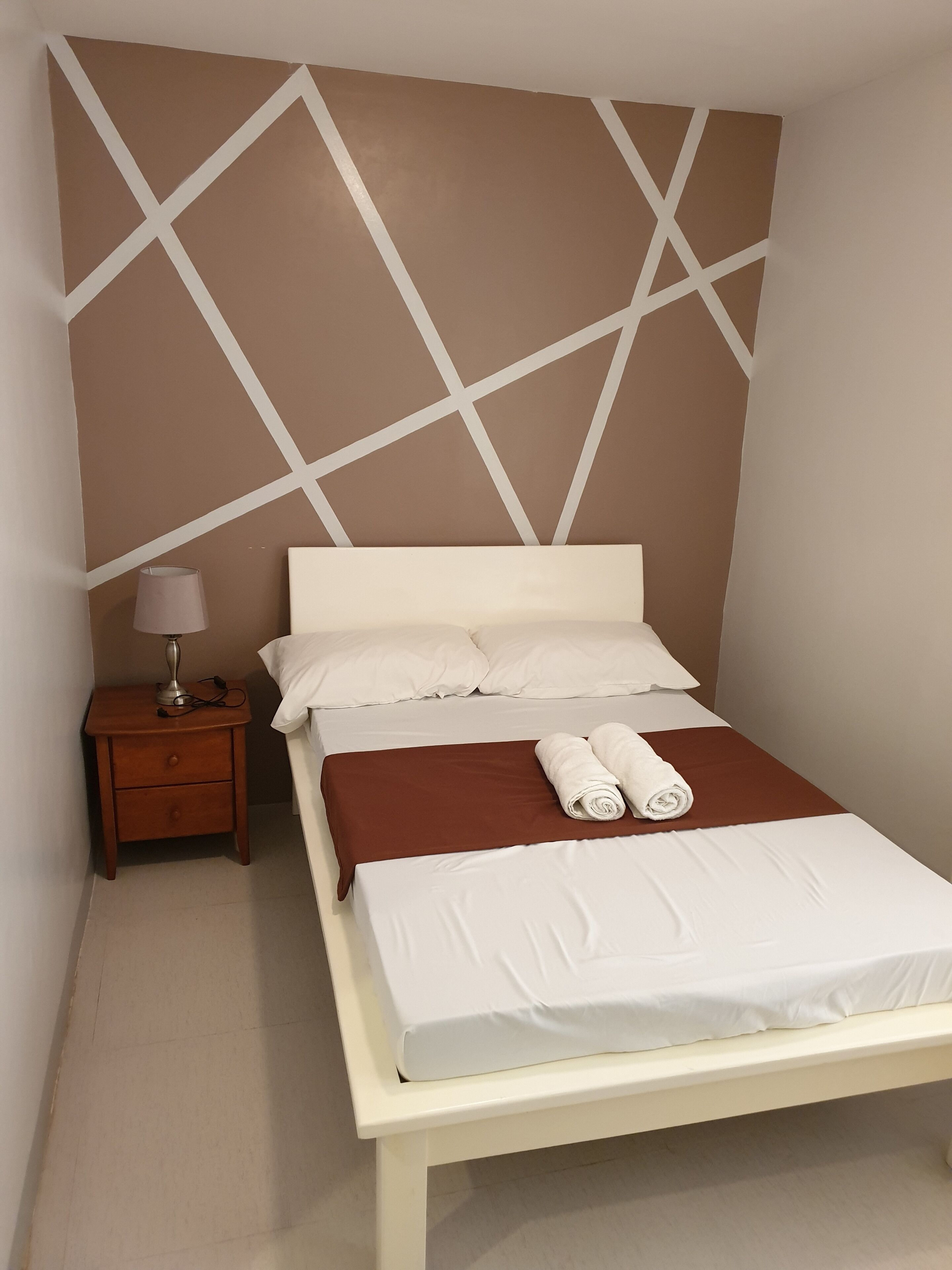 Una cama bien hecha con sábanas blancas y un plaid marrón, una mesita de noche de madera con una lámpara, y una pared con un diseño geométrico.