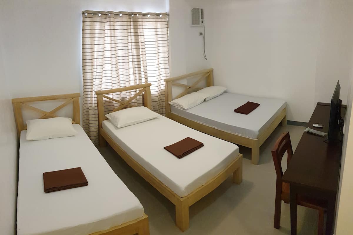 A room with three wooden beds, each with a white mattress and a brown pillow. There is a wooden desk with a chair, a television, and a window with curtains.