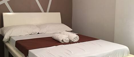 A single bed with white linens, a brown blanket, and two rolled towels.