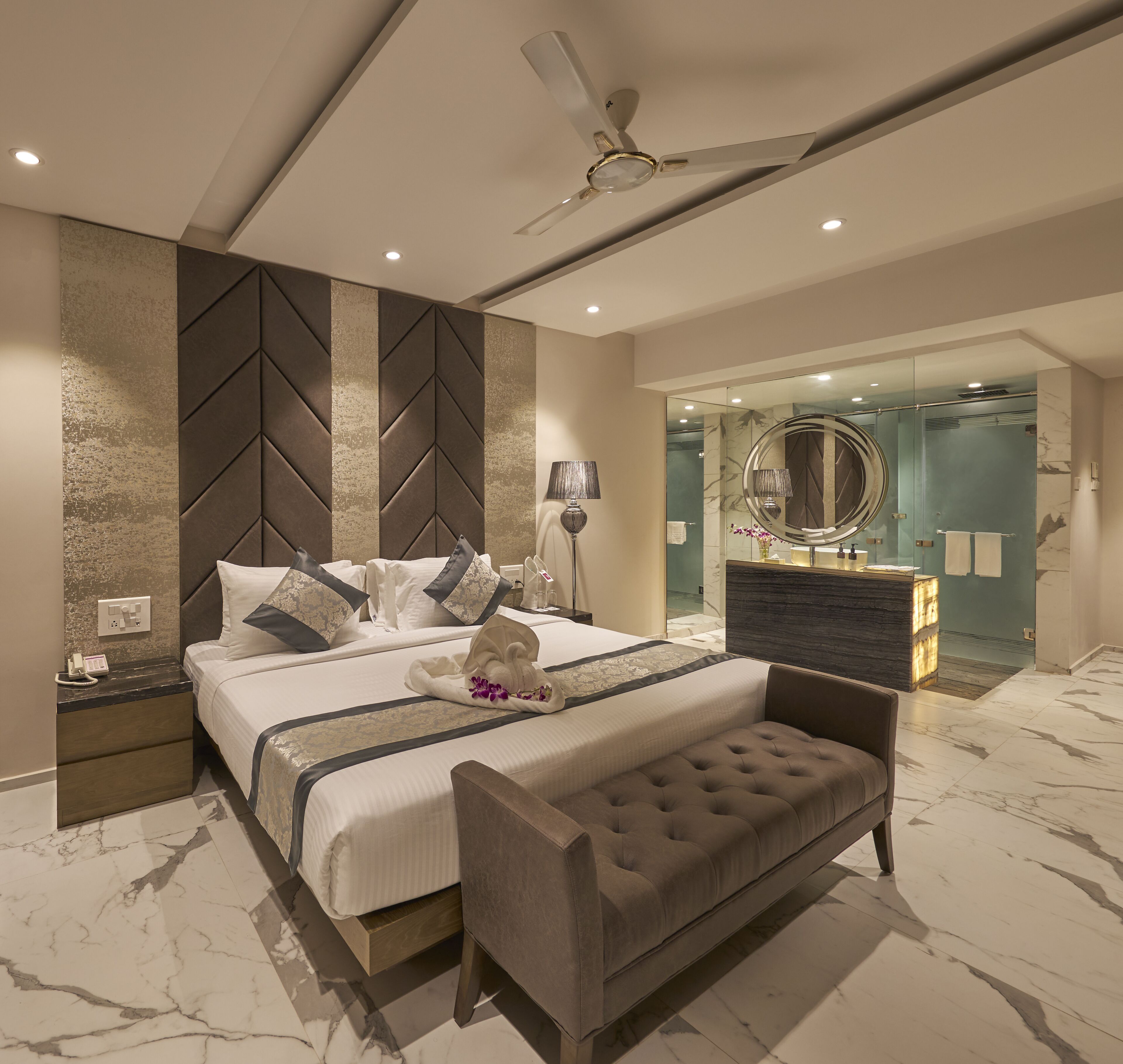 A modern bedroom with a bed, bedside tables, a bench, a ceiling fan, and a bathroom visible through a glass door.