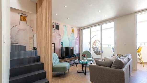 Un salon moderne avec un canapé, un téléviseur et un coin repas avec des chaises. Un escalier mène à un autre niveau.