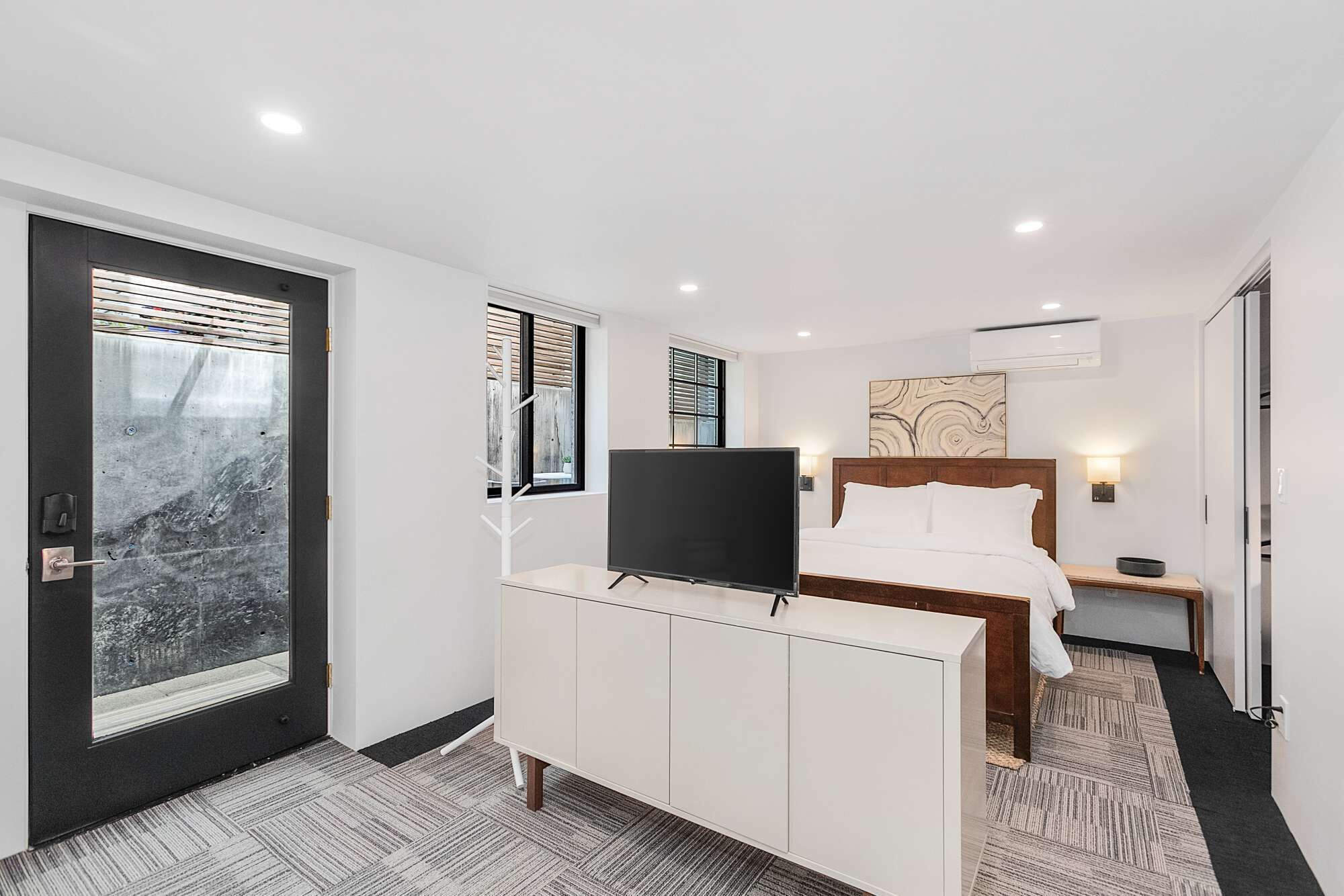 A hotel room with a bed, a TV, a desk, and a door with a view of the outside.