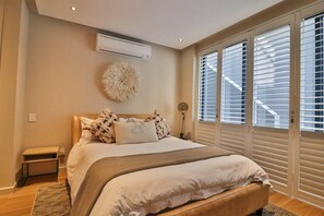 A bedroom with a large bed, a small bedside table, a wall-mounted air conditioner, and a large window with blinds.