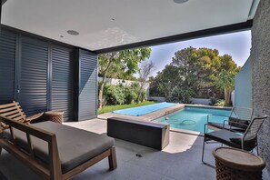 A modern outdoor patio with a pool, wooden furniture, and a view of the surrounding greenery.
