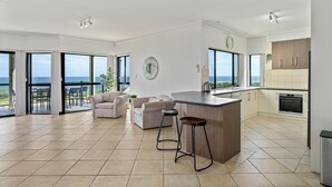 Een ruime woonkamer met een groot raam met uitzicht op zee, een keuken met moderne apparatuur, en een gezellige zithoek met twee banken en stoelen.