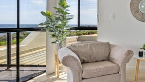 Een ruime kamer met grote ramen, een beige fauteuil en een potplant.