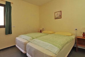 A double bed with green and white bedding, a small bedside table with a lamp, a window with a green curtain, and a painting on the wall. - Bungalow Moijama 1, Puerto Naos (Puerto Naos)