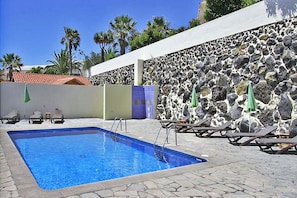 A swimming pool with lounge chairs and umbrellas, surrounded by a stone wall and palm trees. - Bungalow Moijama 1, Puerto Naos (Puerto Naos)
