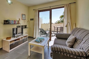 A living room with a sofa, a TV on a stand, a small table, and a balcony with a chair. - Bungalow Moijama 1, Puerto Naos (Puerto Naos)