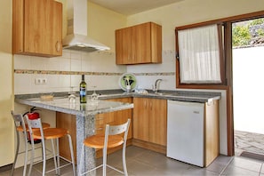 A compact kitchen with wooden cabinets, a granite countertop, a small refrigerator, and two bar stools. - Bungalow Moijama 1, Puerto Naos (Puerto Naos)
