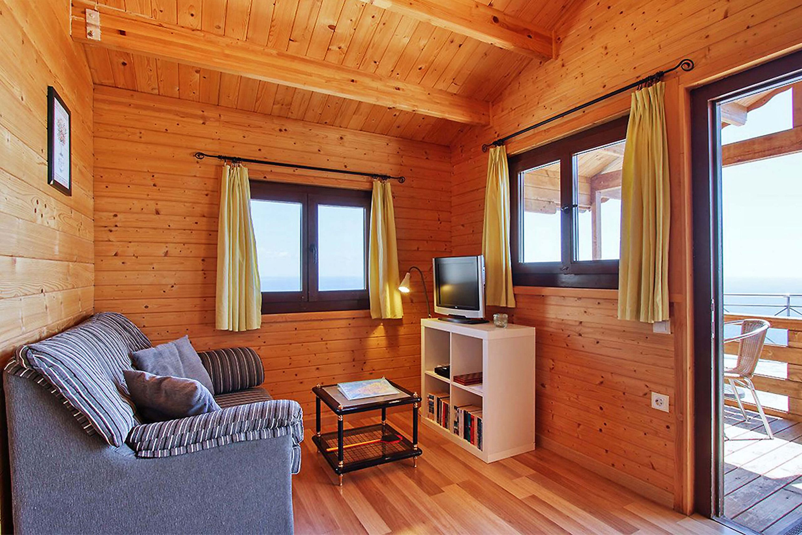 Interior acolhedor de uma cabana com sofá, uma mesa pequena, TV e uma varanda com uma cadeira.