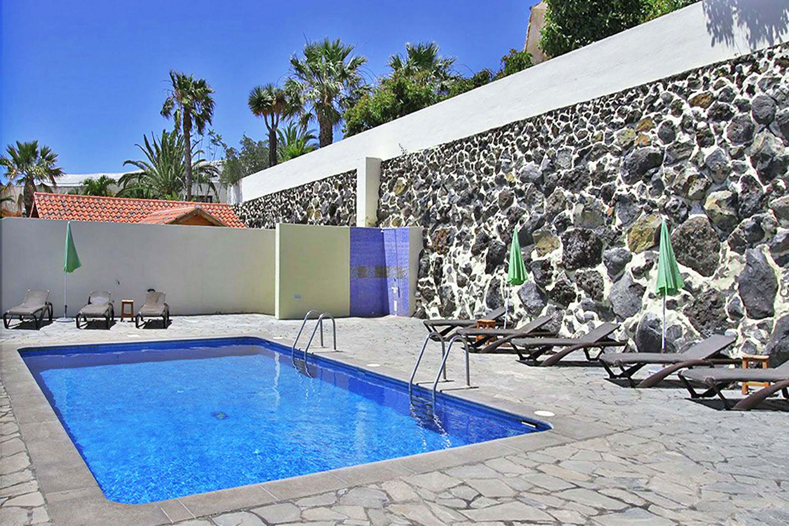 Une piscine entourée d’un mur de pierre et de palmiers, avec des chaises longues et des parasols.