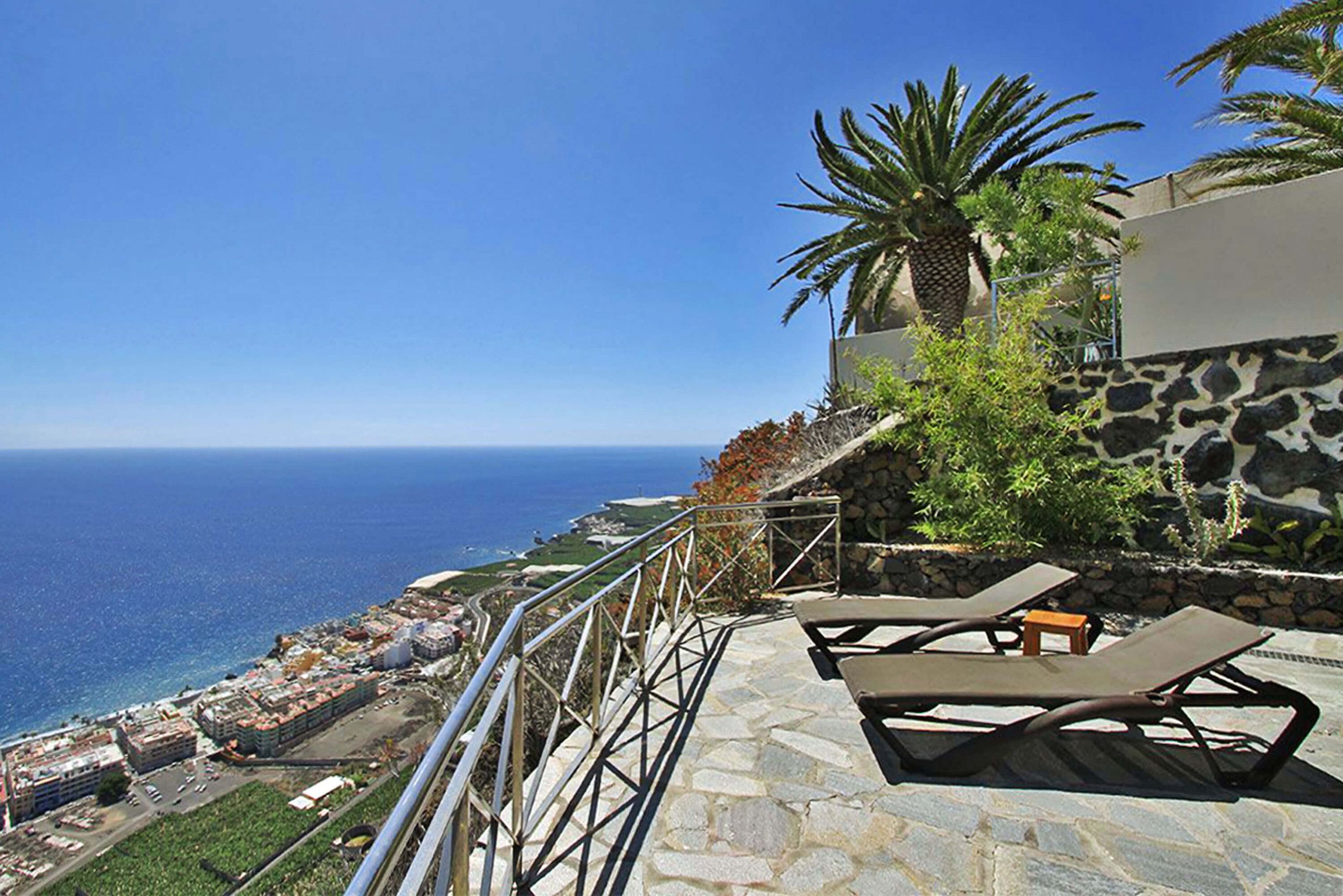 Une terrasse en bord de mer, dotée de chaises longues, d’un mur en pierre et offrant une vue sur l’océan.