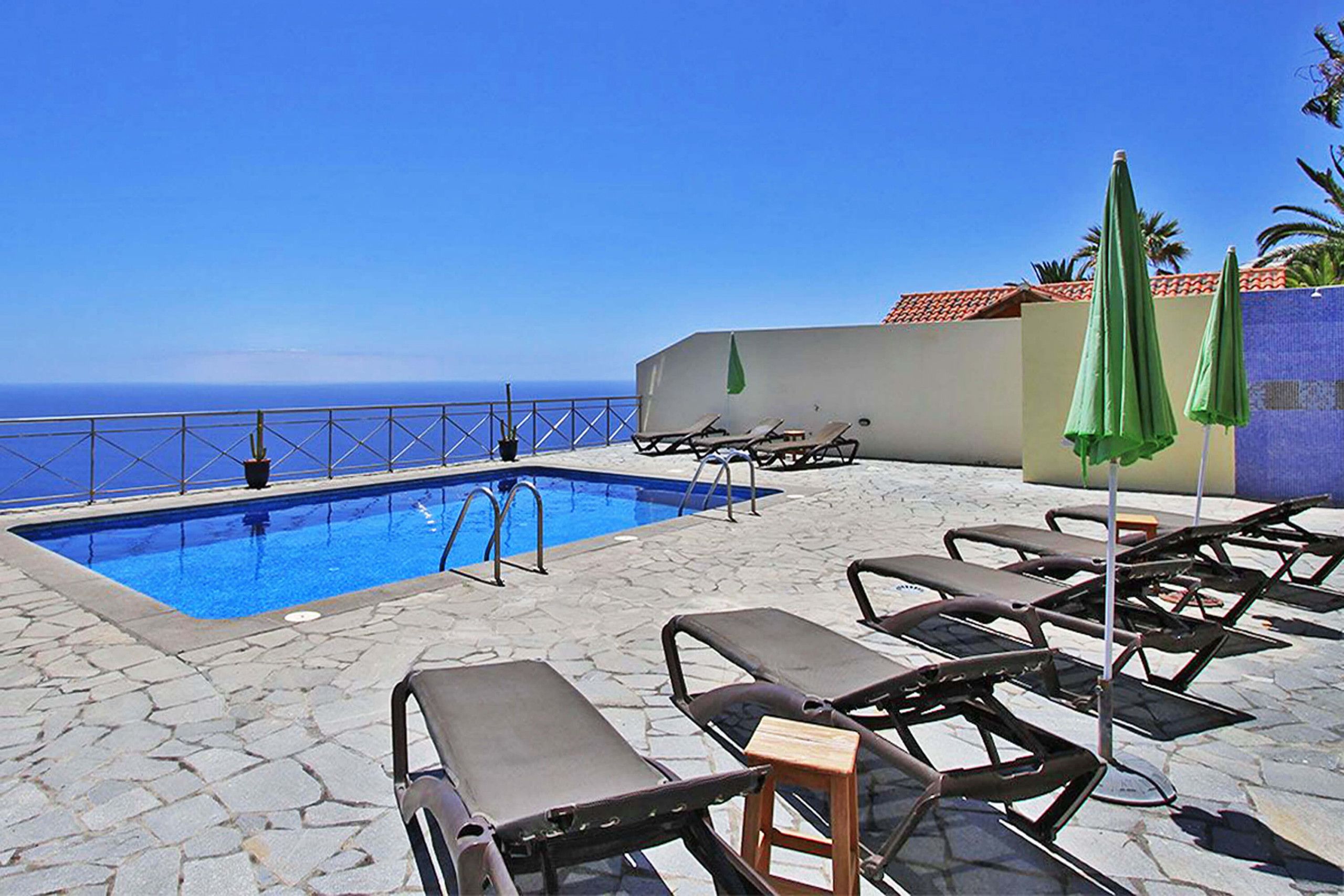 Uma área de piscina com espreguiçadeiras e guarda-sóis, céu azul límpido e vista para o oceano.