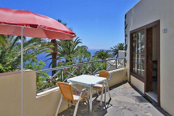 Ein Balkon mit Tisch und Stühlen, einem roten Sonnenschirm und Blick auf das Meer.