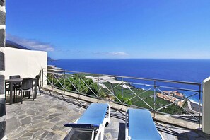 A terrace with stone flooring, two blue sun loungers, a table with chairs, and a view of the ocean and distant buildings. - Bungalow Moijama 8, Puerto Naos (Puerto Naos)