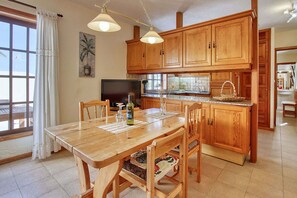 Une cuisine avec des meubles de rangement en bois, une table à manger avec des chaises, un téléviseur et une fenêtre avec des rideaux.
