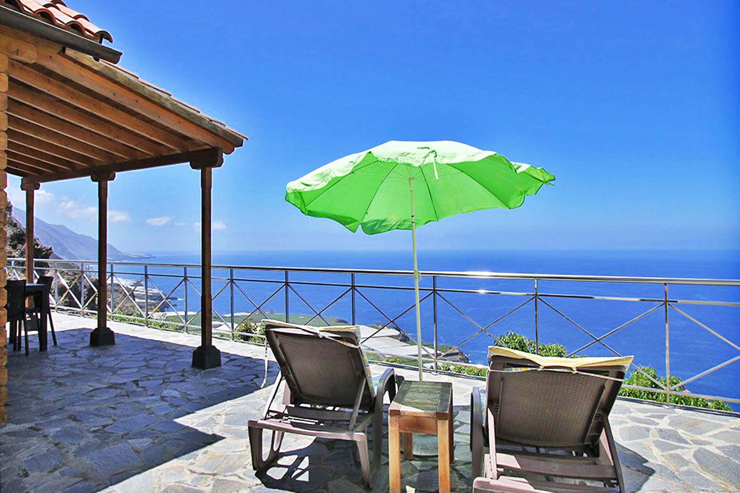 Un espacio al aire libre con una sombrilla verde, suelo de piedra y vistas al mar.