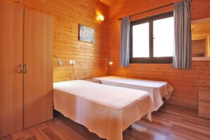 A wooden cabin room with two beds, a wardrobe, a window with curtains, and a wall-mounted light. - Bungalow Moijama 12, Puerto Naos (Puerto Naos)