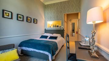 A hotel room with a bed, a desk with a lamp, a chair, and framed pictures on the wall.