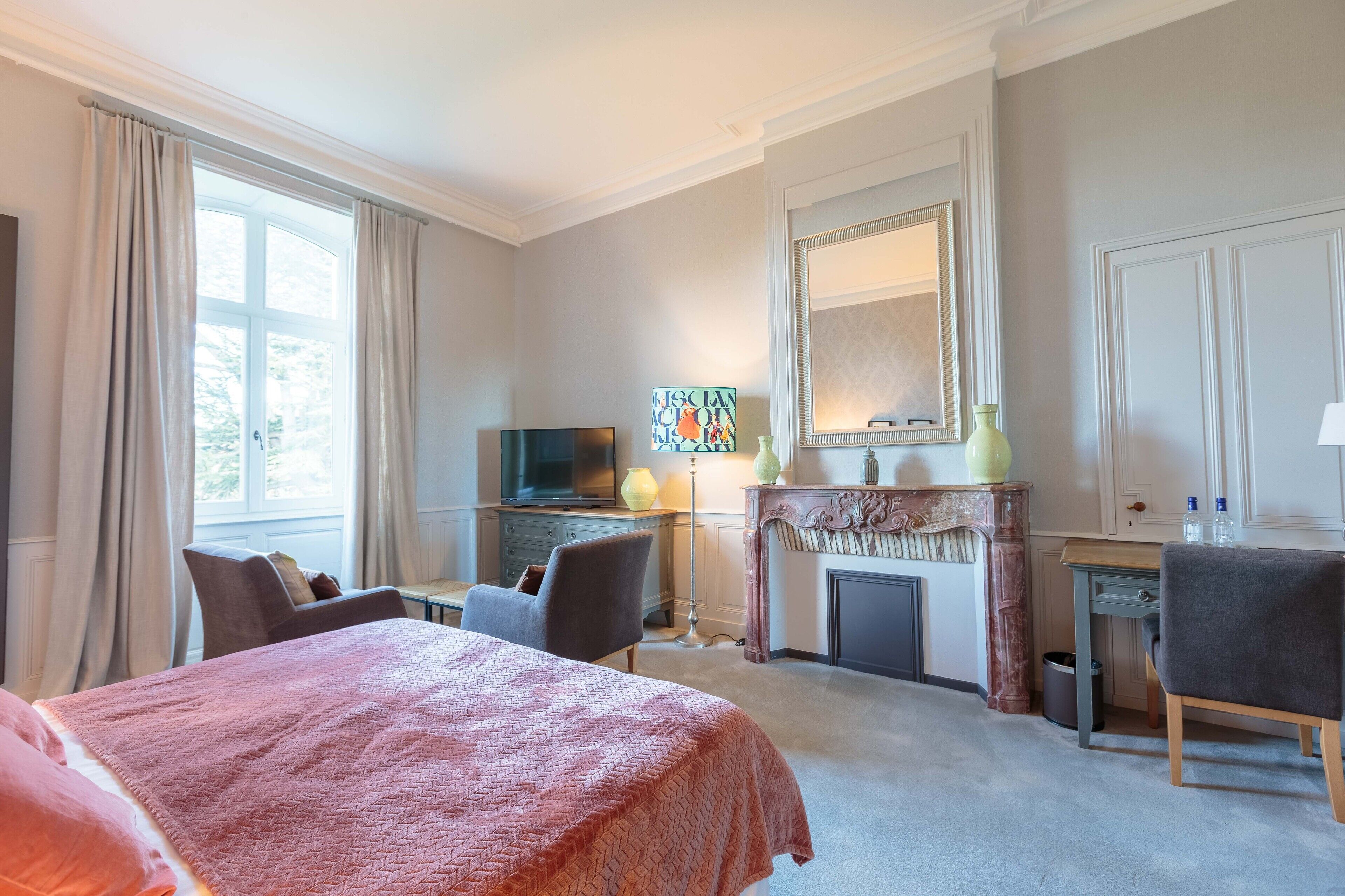 Habitación de hotel con una cama grande, un escritorio con silla, televisor, chimenea y un ventanal con cortinas.