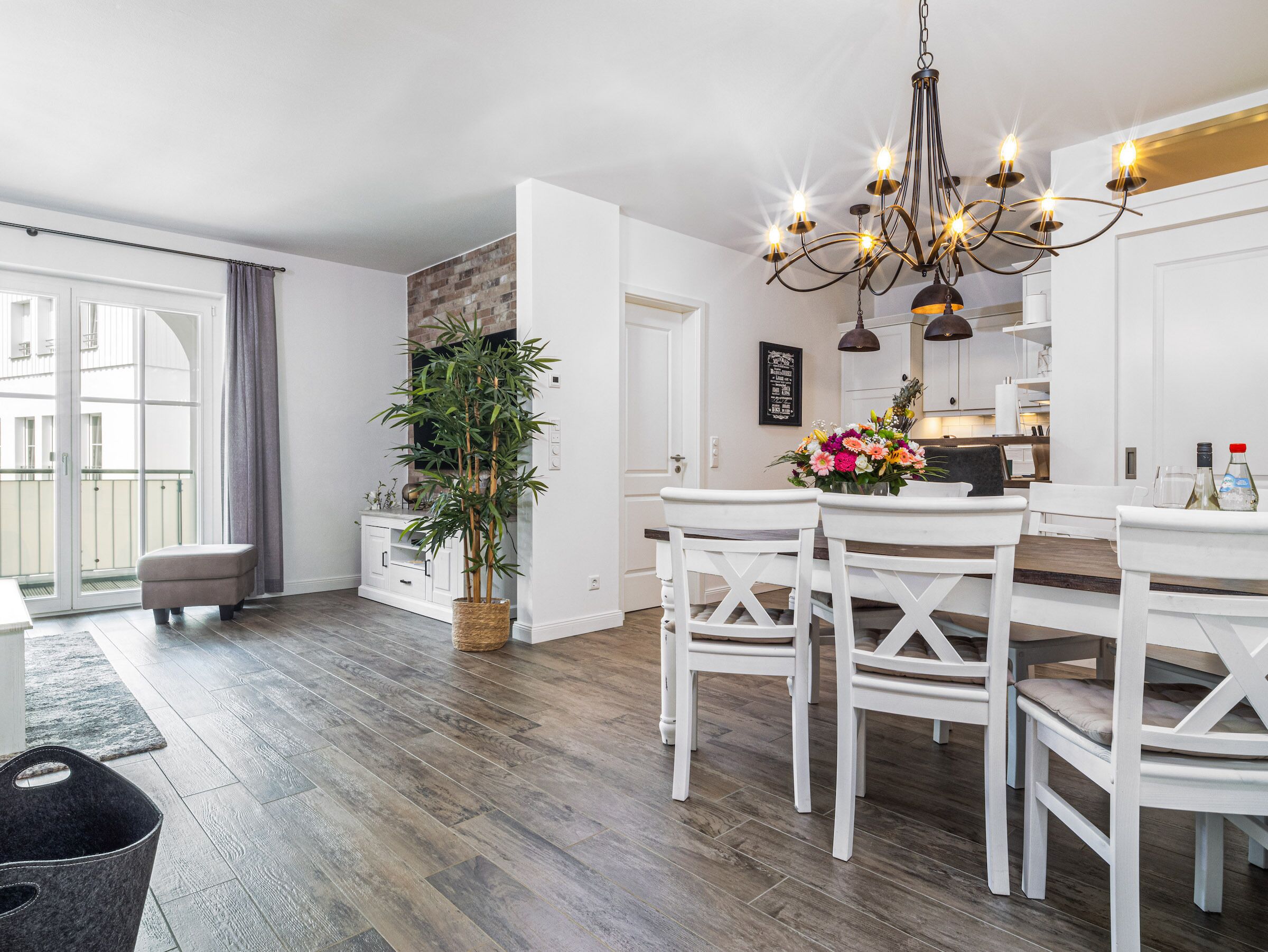 A modern kitchen with white cabinetry, a dining area with white chairs, a chandelier, and a potted plant.