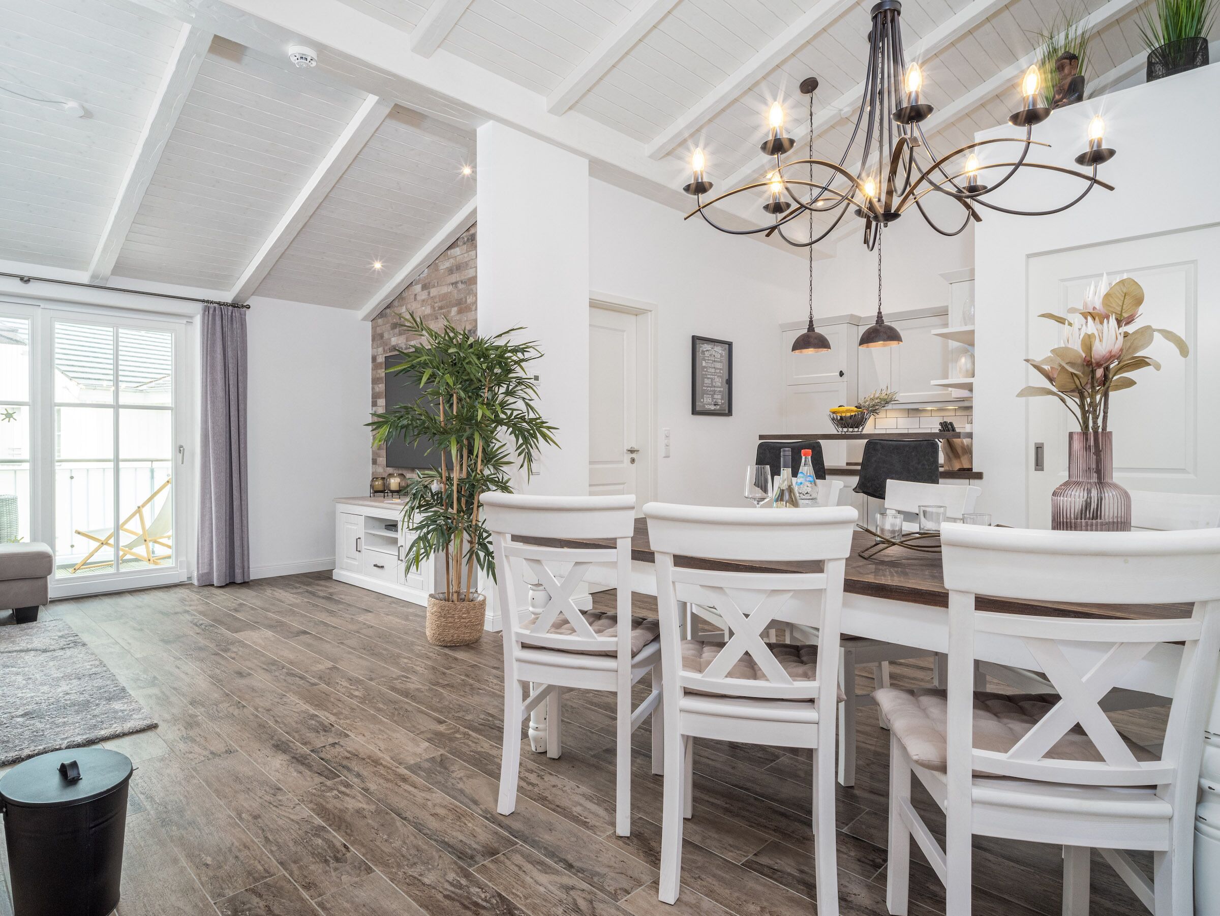 A spacious dining area with a high ceiling, white walls, and a wooden floor. There is a chandelier hanging above the table, and a potted plant is visible in the background.