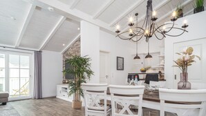 Un'ampia sala da pranzo con soffitto alto, pareti bianche e pavimento in legno. Al di sopra del tavolo è appesa una lampadario, e sullo sfondo si intravede una pianta in vaso.