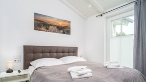 Un letto rifatto con precisione, con una coperta grigia e cuscini bianchi, un comodino con una lampada, una presa elettrica e un quadro appeso alla parete raffigurante un tramonto sulla spiaggia.