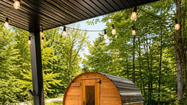 A wooden barrel sauna with a door, surrounded by a covered patio with two green lounge chairs. - New Family Cottage Tremblant-4 Bdrs w/ Hot Tub (La Conception)