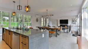 A modern kitchen with a large island, pendant lighting, and large windows offering a view of the outdoors. - New Family Cottage Tremblant-4 Bdrs w/ Hot Tub (La Conception)