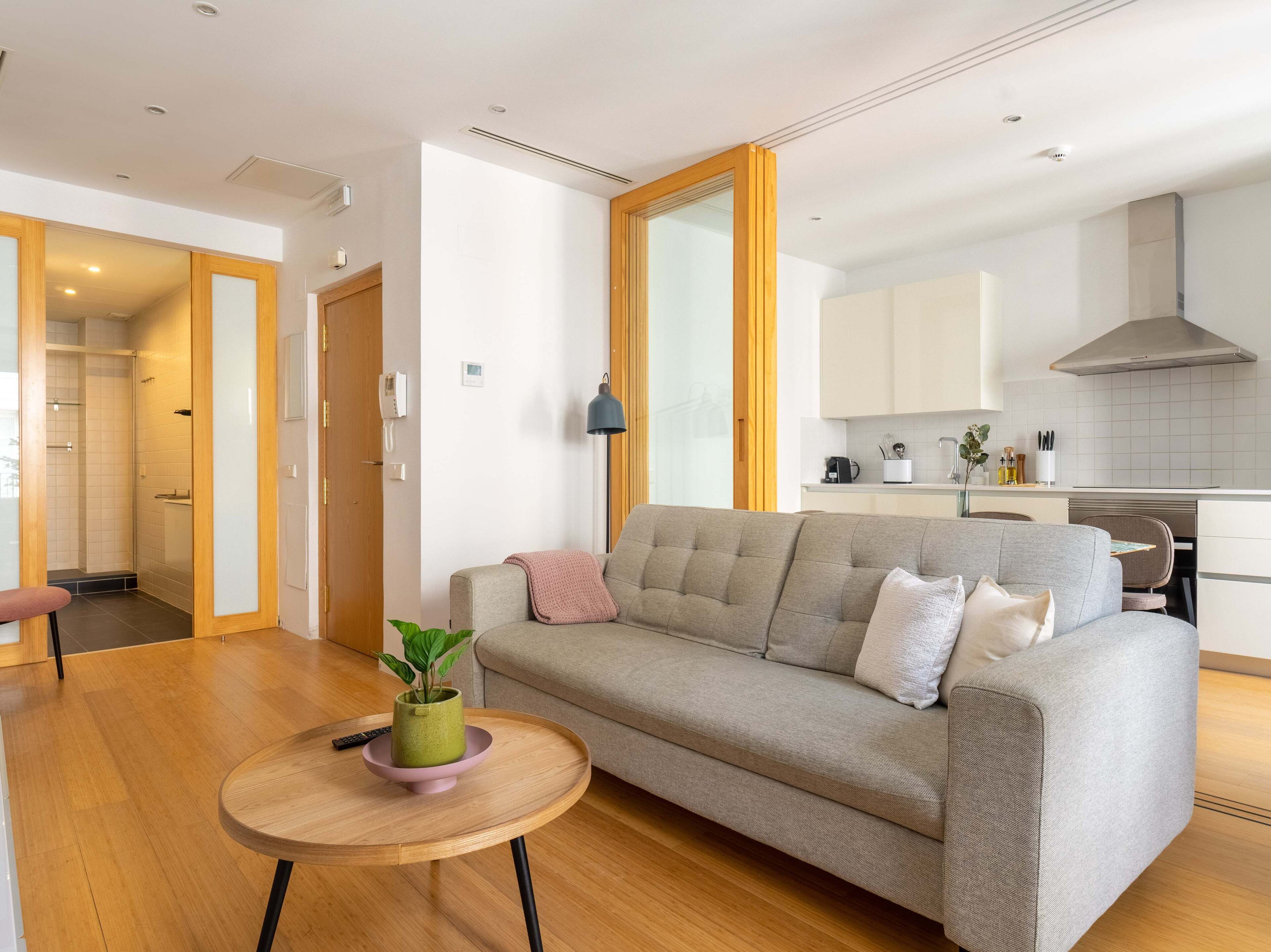 Ein modernes Wohnzimmer mit einem grauen Sofa, einem runden Holztisch mit einer Zimmerpflanze und einer Küche im Hintergrund.