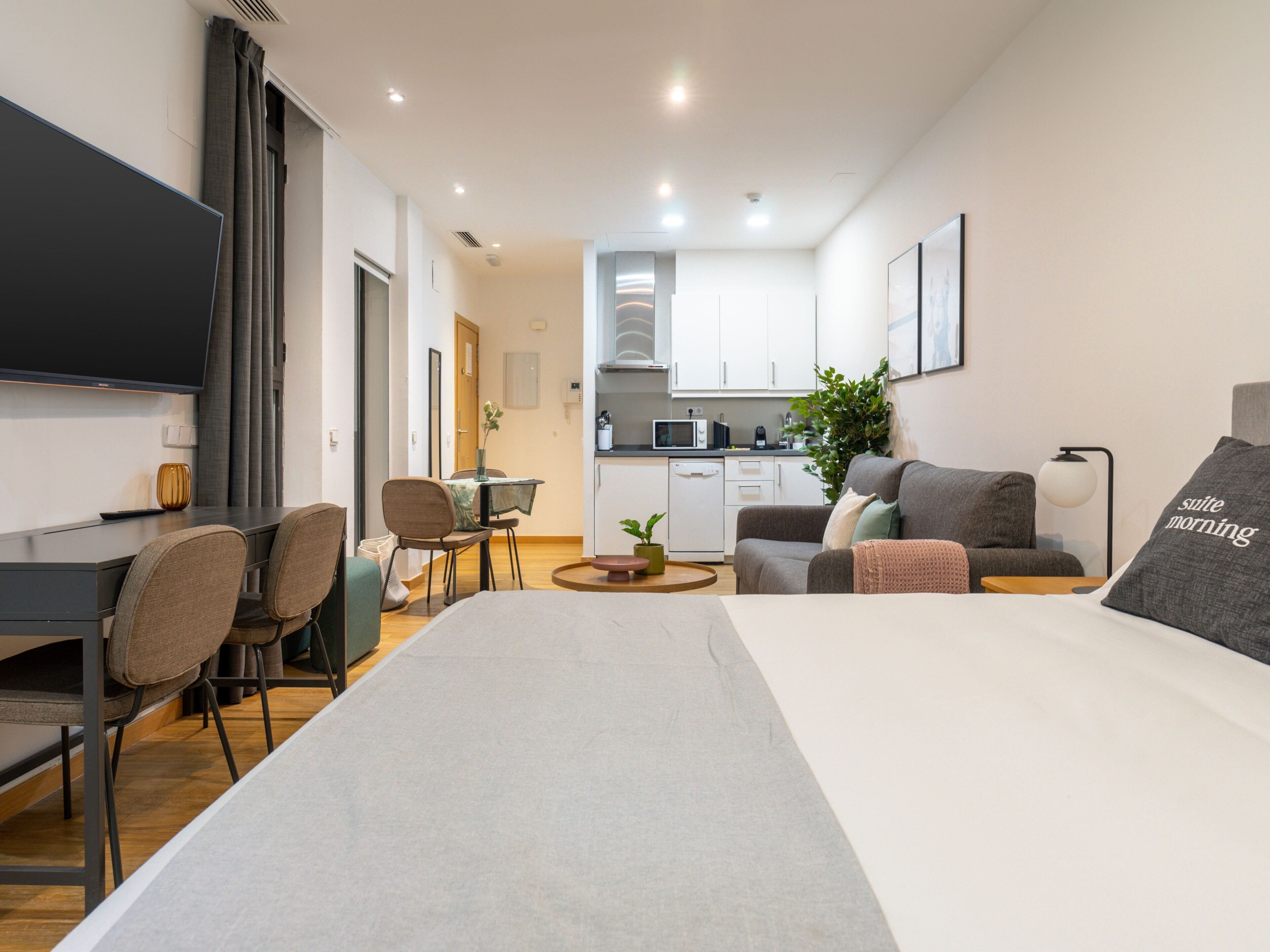A modern hotel room with a flat-screen TV, a dining area with chairs and a table, a kitchenette with a microwave, and a sofa with a pillow that reads "suite morning".
