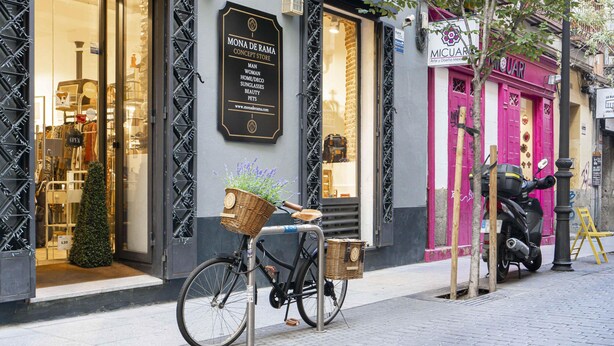 Een etalage met een bordje voor 'Mona de Rama Concept Store' en een fiets met een mandje op het trottoir.