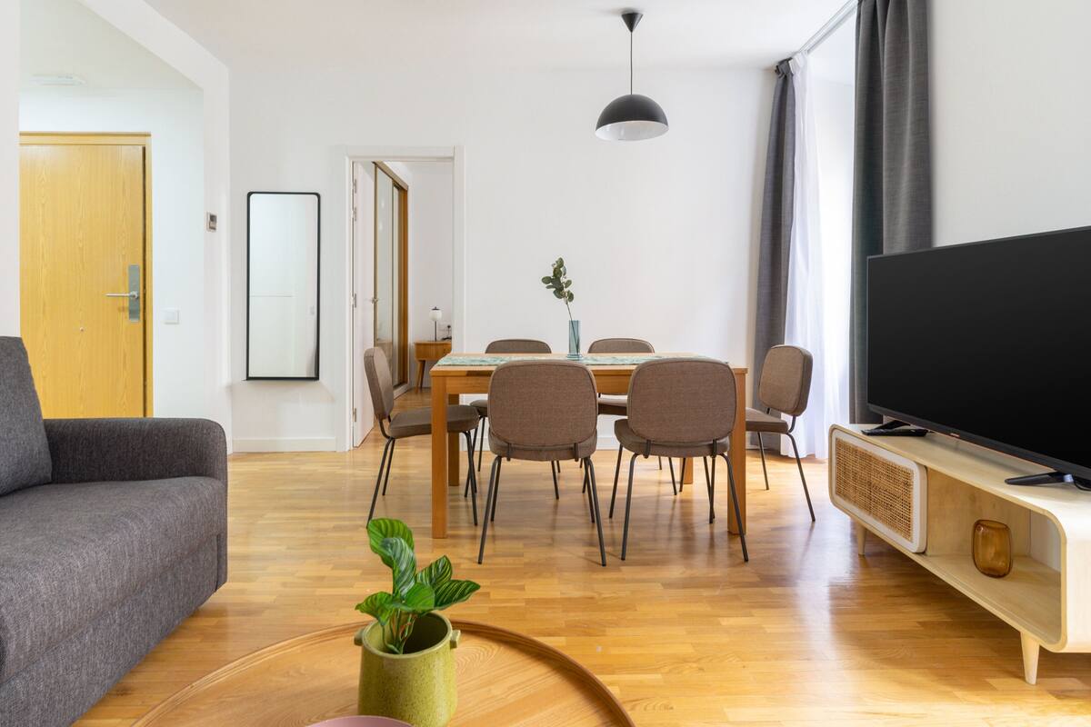 Ein modernes Wohnzimmer mit einer Couch, einem Esstisch, Stühlen, einem Fernseher und einer Topfpflanze.