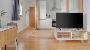 Een moderne woonkamer met een flatscreen televisie op een houten standaard, een eethoek met een tafel en stoelen, en een keuken op de achtergrond.