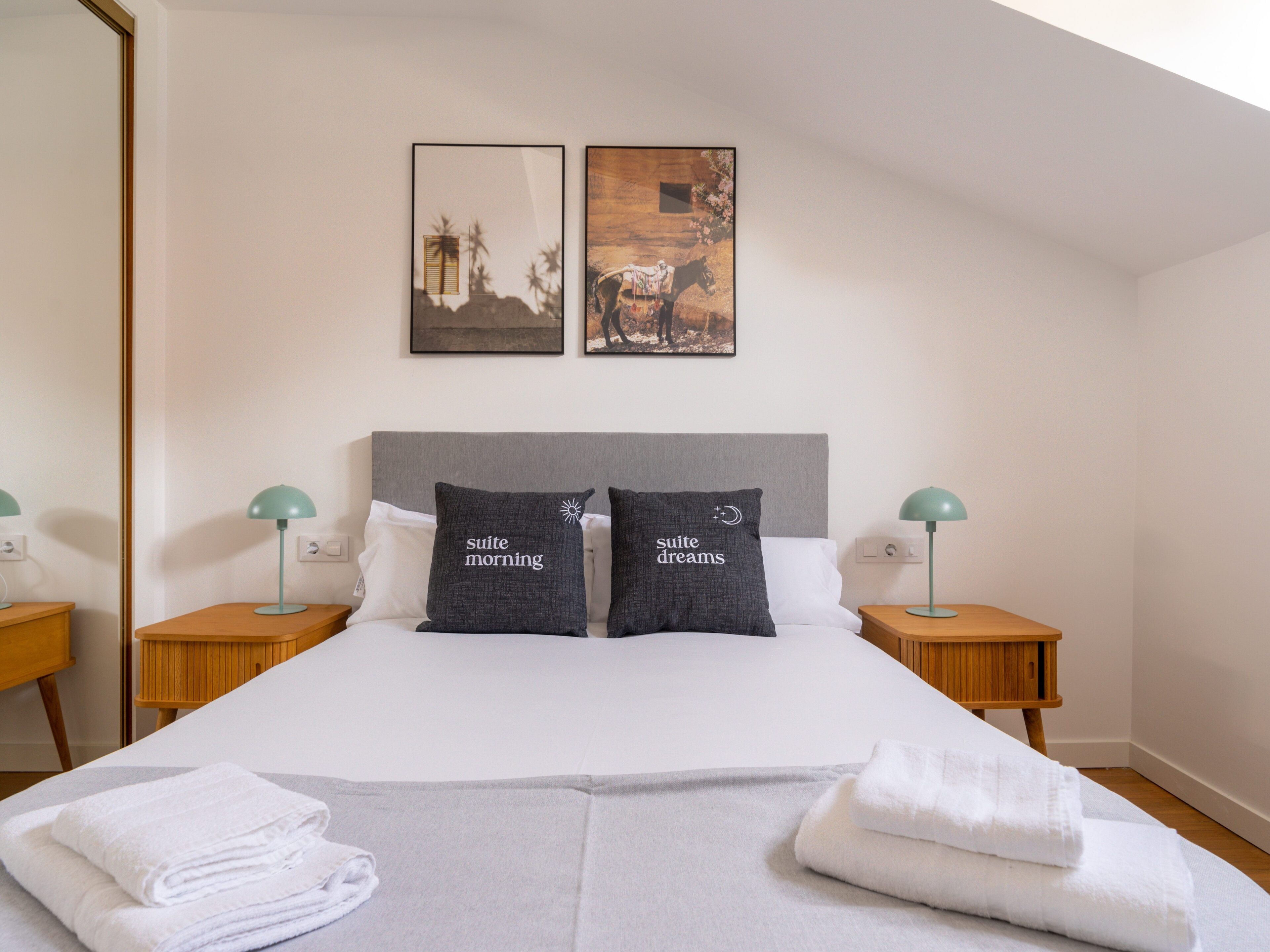 A neatly made bed with two pillows, a bedside table with a lamp, and two framed pictures on the wall.