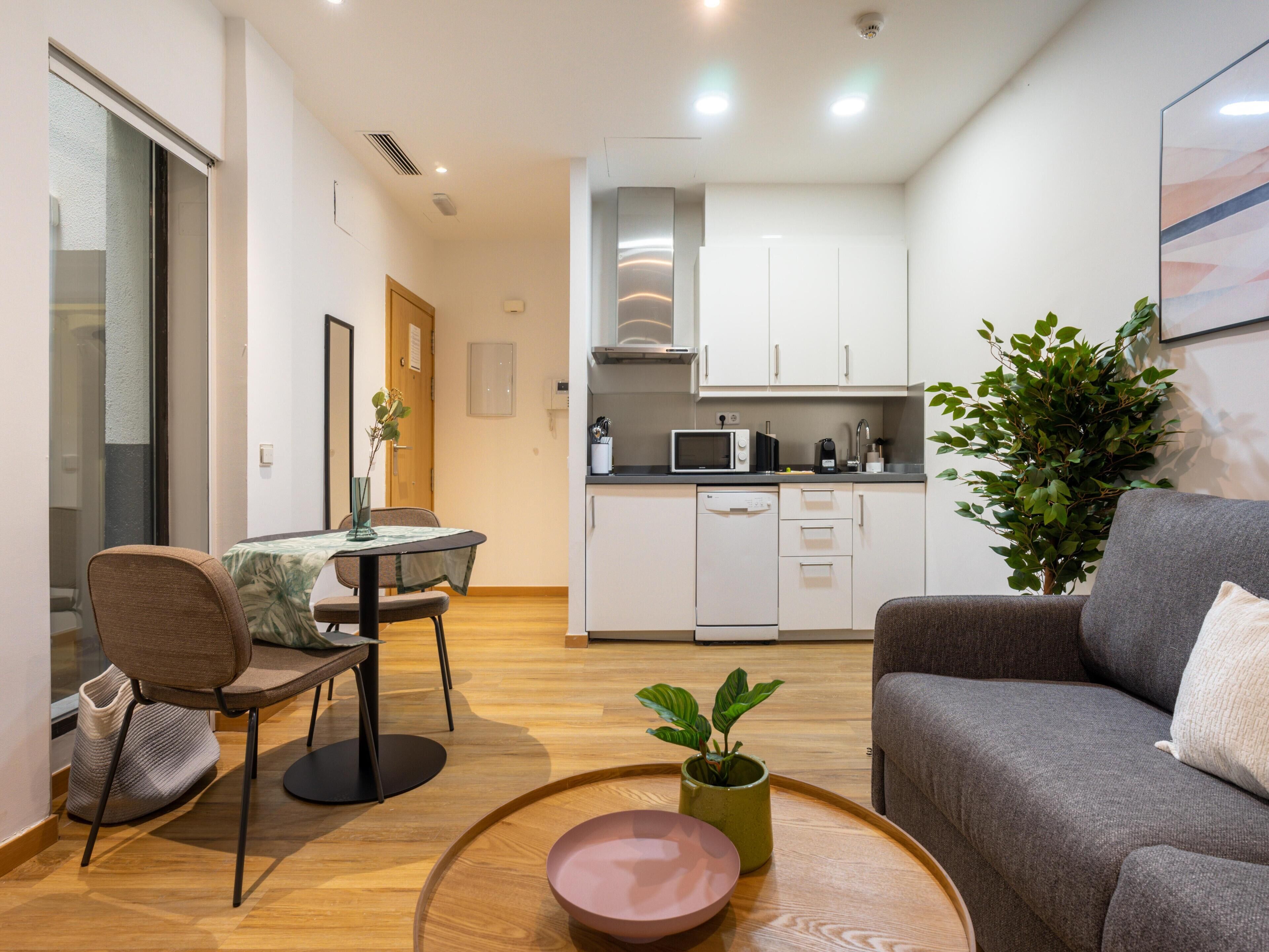 Un salon moderne avec un canapé gris, une table à manger avec des chaises et un coin cuisine doté de meubles blancs et d’appareils électroménagers.