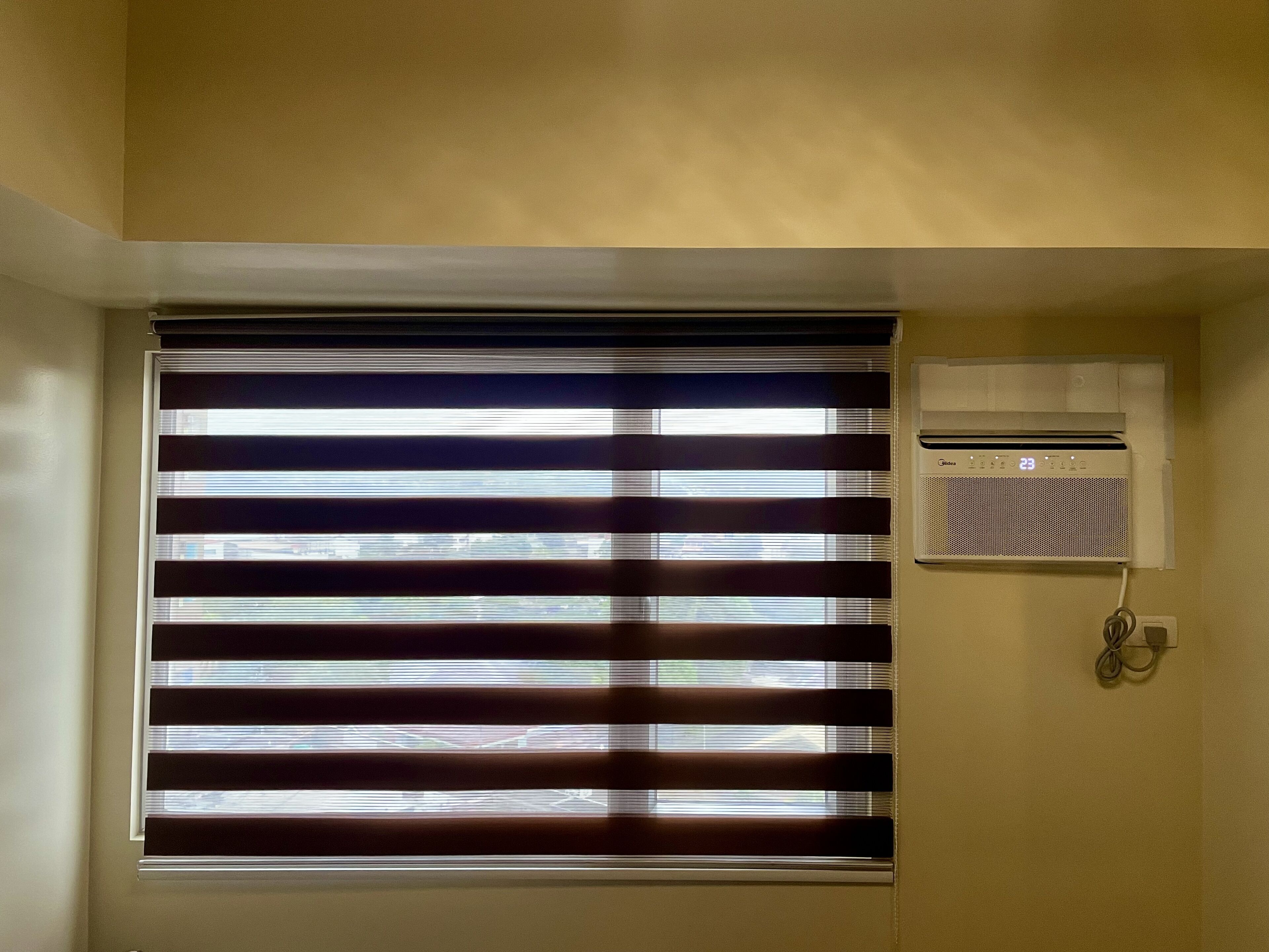A room with horizontal blinds, a wall-mounted air conditioner, and a visible power outlet.