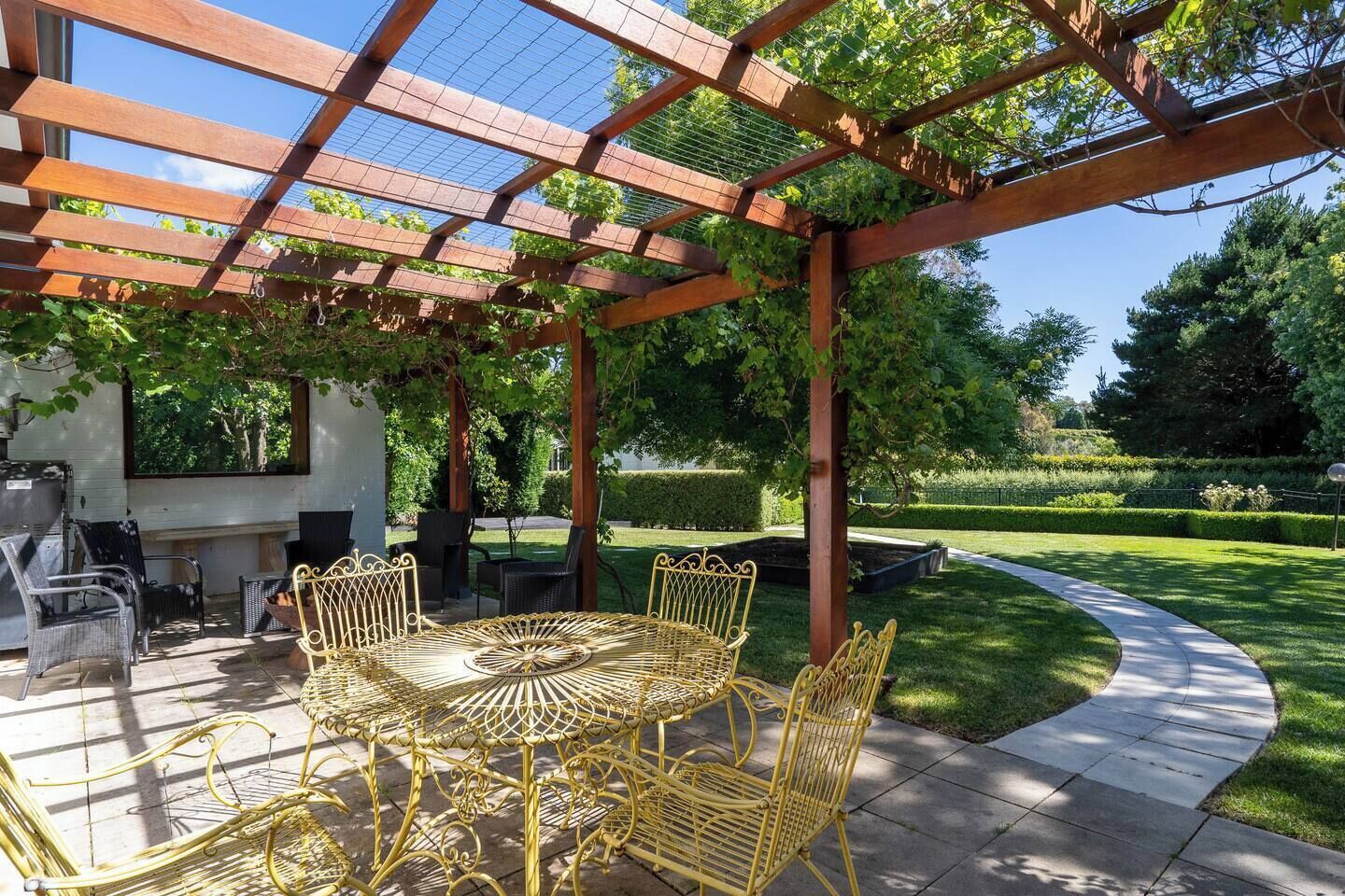 A covered outdoor patio with a round table and chairs, surrounded by greenery and a well-maintained lawn.