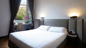 Ein Hotelzimmer mit einem großen Bett, einem Schreibtisch mit Stuhl, einem Nachttischtelefon und einem Vorhangfenster.