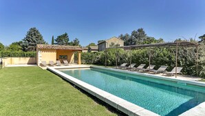 Ein rechteckiger Pool mit Fliesenrand, umgeben von einer gepflegten Rasenfläche. Entlang des Poolrandes sind Liegestühle aufgereiht, und im Hintergrund ist eine Pergola mit Dach zu sehen.