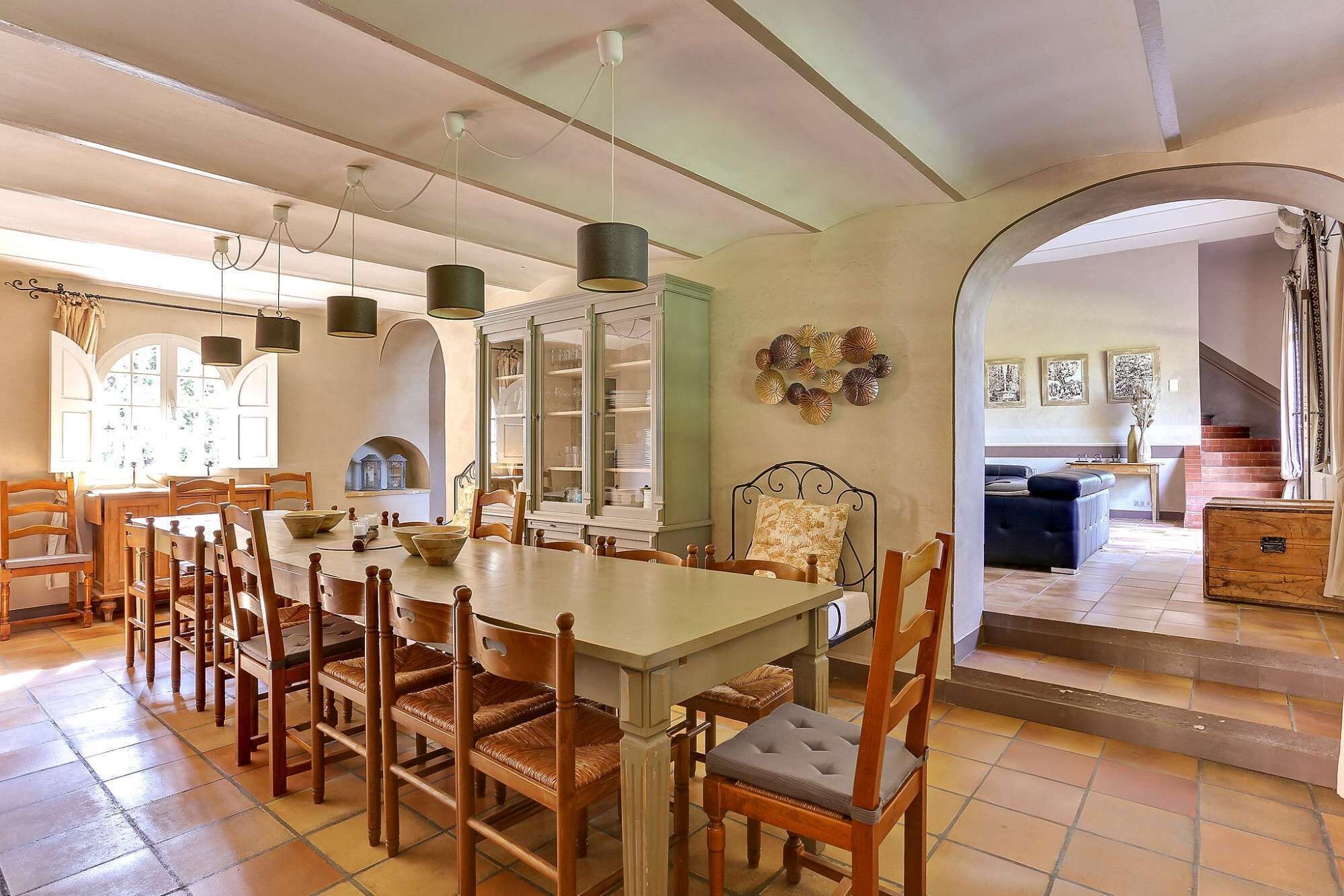 Un espace repas avec une grande table et plusieurs chaises, une cuisine visible à travers une porte ouverte, et un escalier menant à un autre niveau.