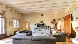 A spacious living room with a large sofa, a coffee table, and a television. There is a fireplace with a wooden mantel and a large mirror above it. The room has a tiled floor and a high ceiling with exposed beams. - Large air-conditioned house with heated swimming pool in quiet Provence (Entraigues sur la Sorgue)